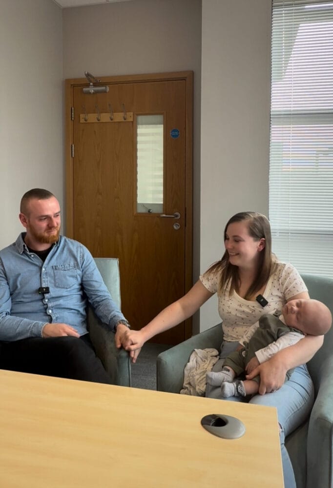 A screenshot of parents Victoria and Jonathan holding hands with their baby on Victoria's lap.